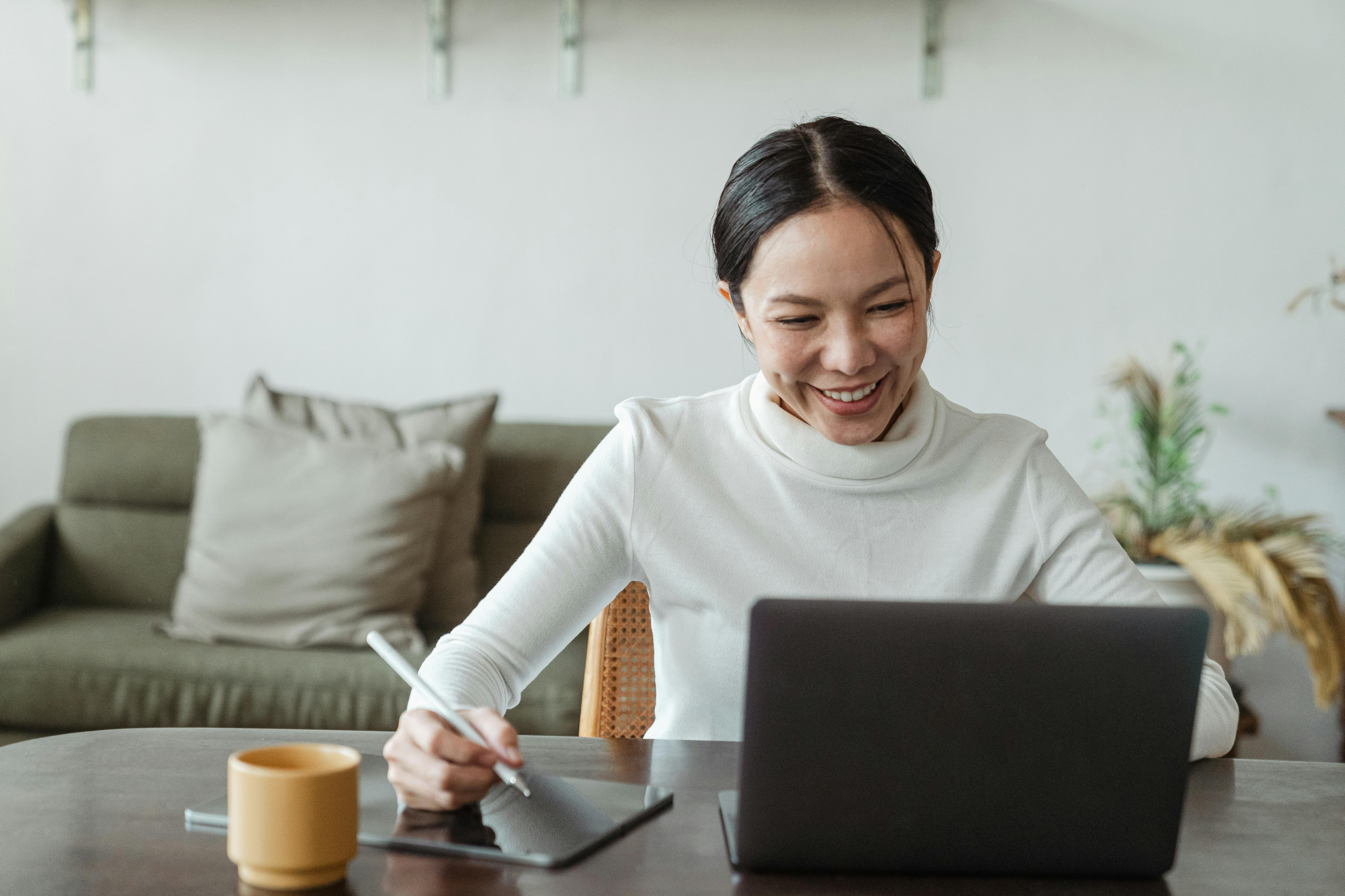 A woman sitting at a table, using a laptop and drawing tablet, smiling