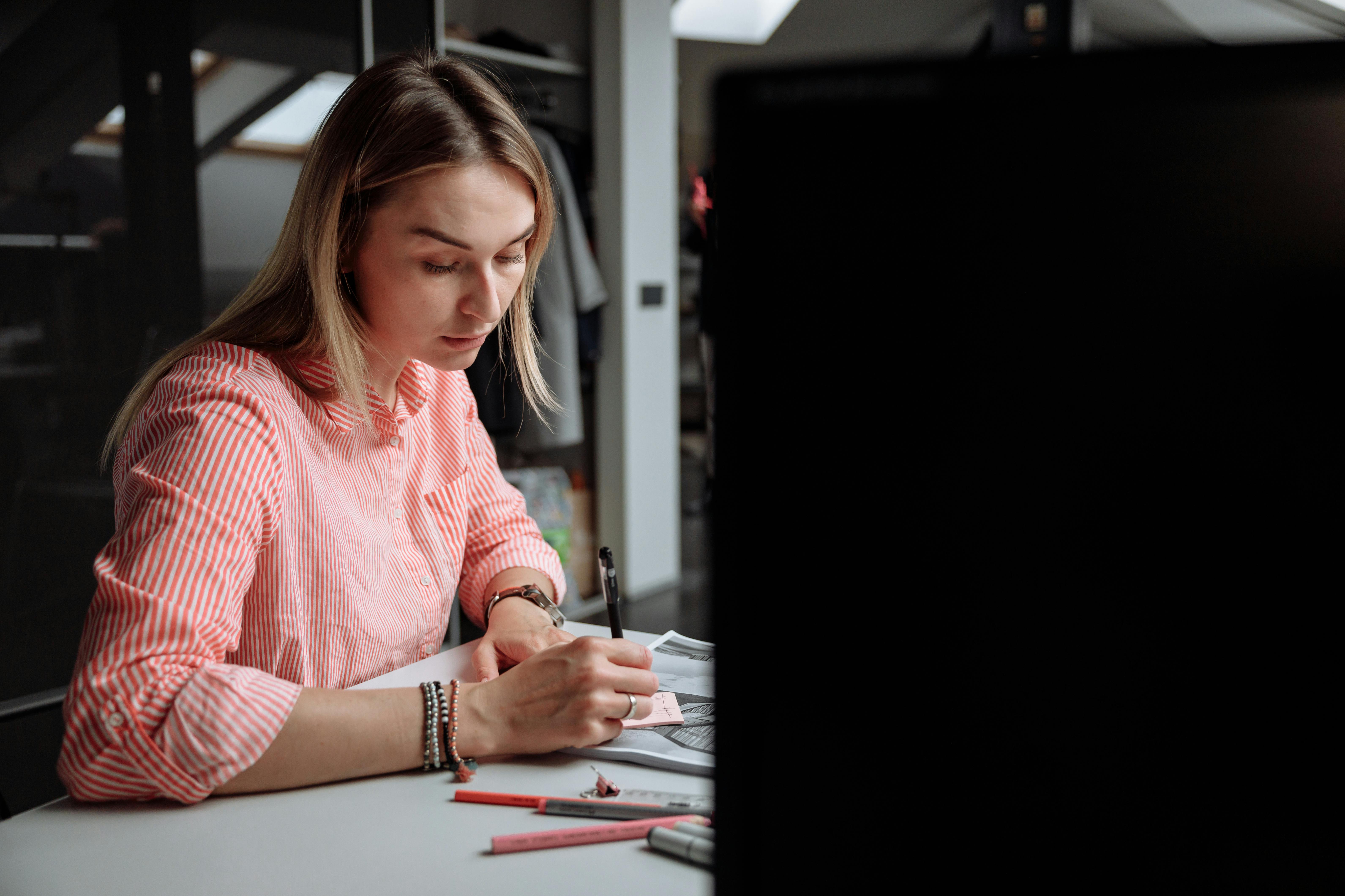 A woman in a pink striped shirt writing at a desk in a studio