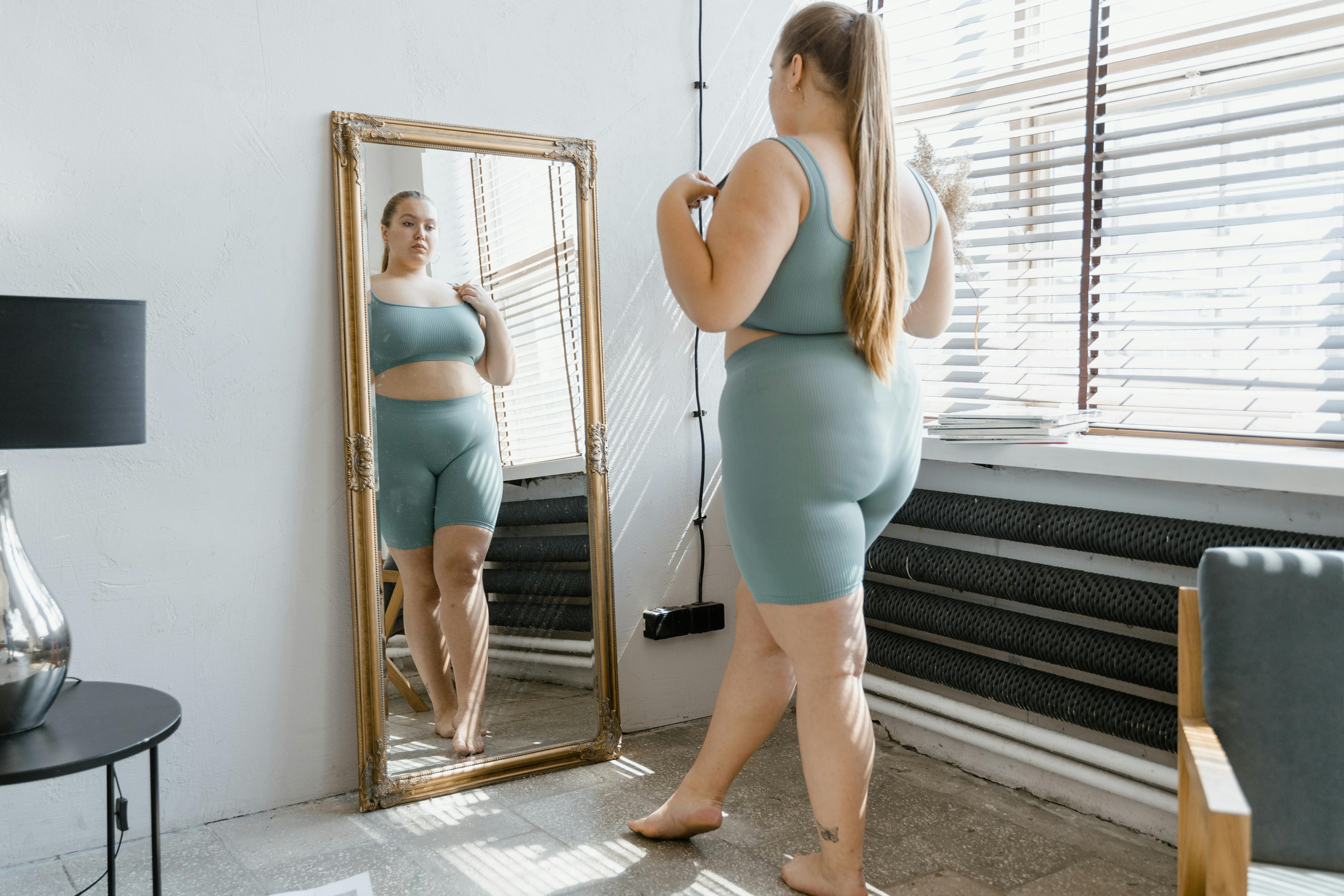 A woman in a teal athletic outfit looking at herself in a mirror.