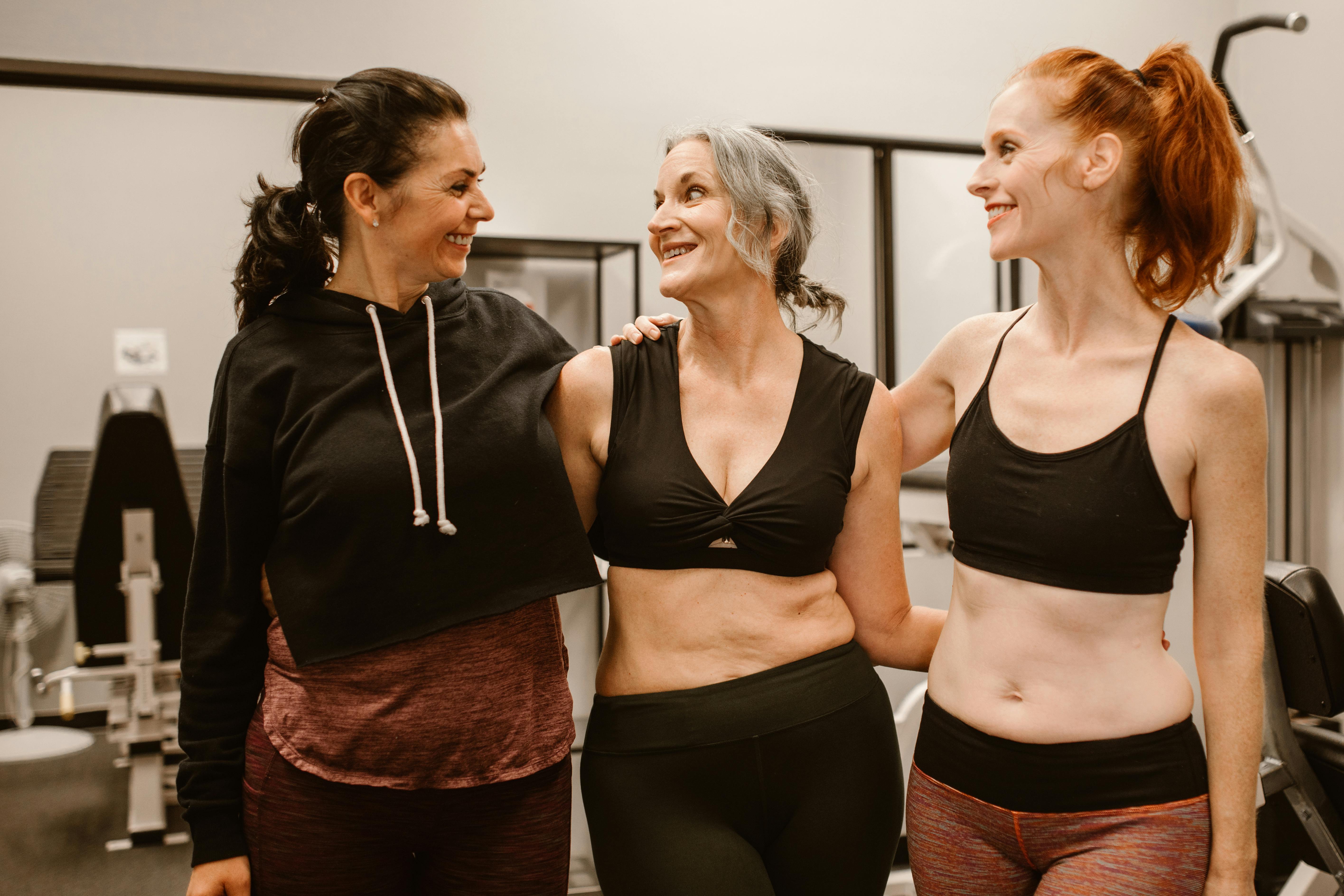 Three women smiling and standing together in a gym environment.