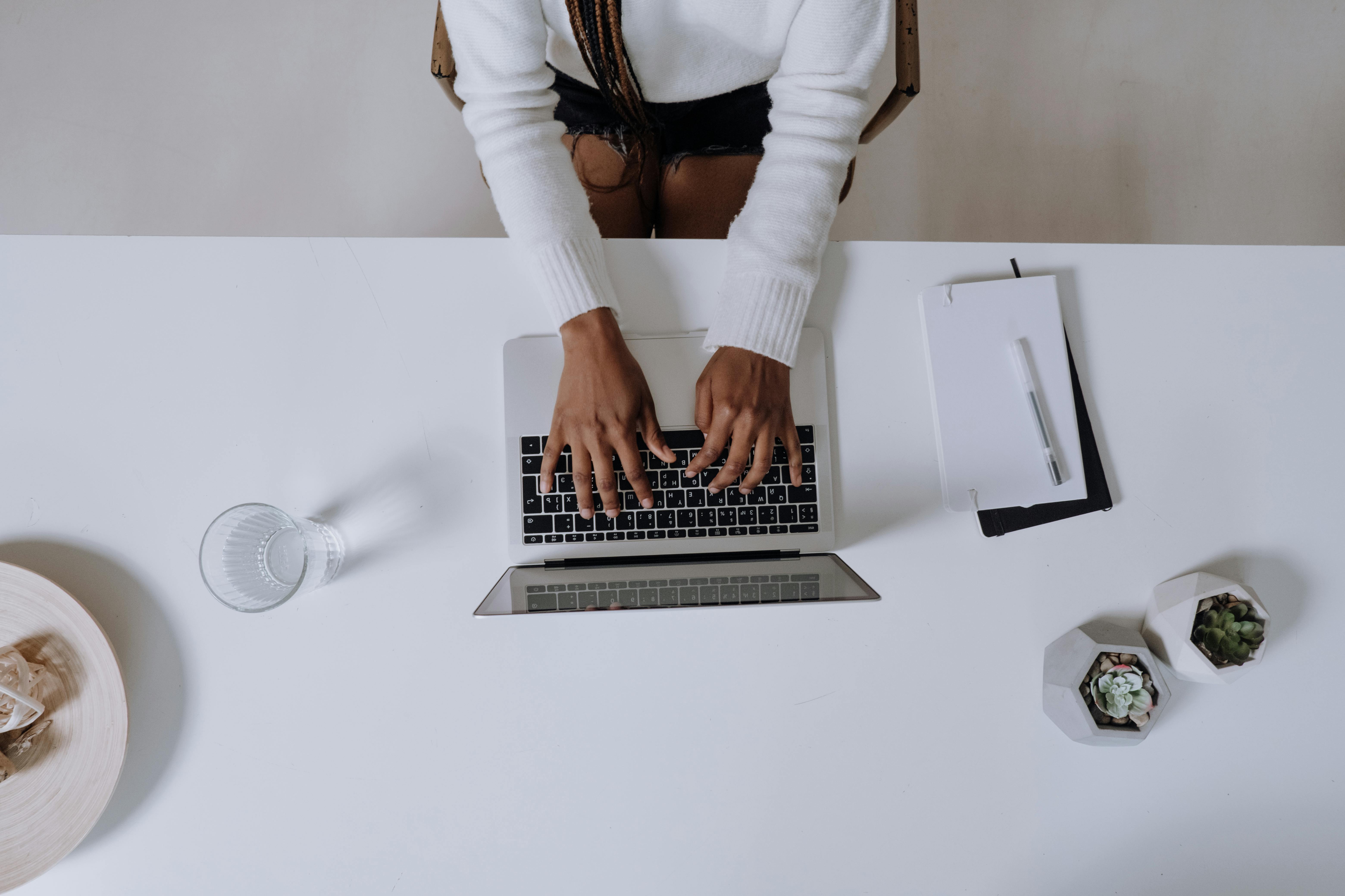 A person typing on a laptop at a white desk, surrounded by a glass of water and potted plants.