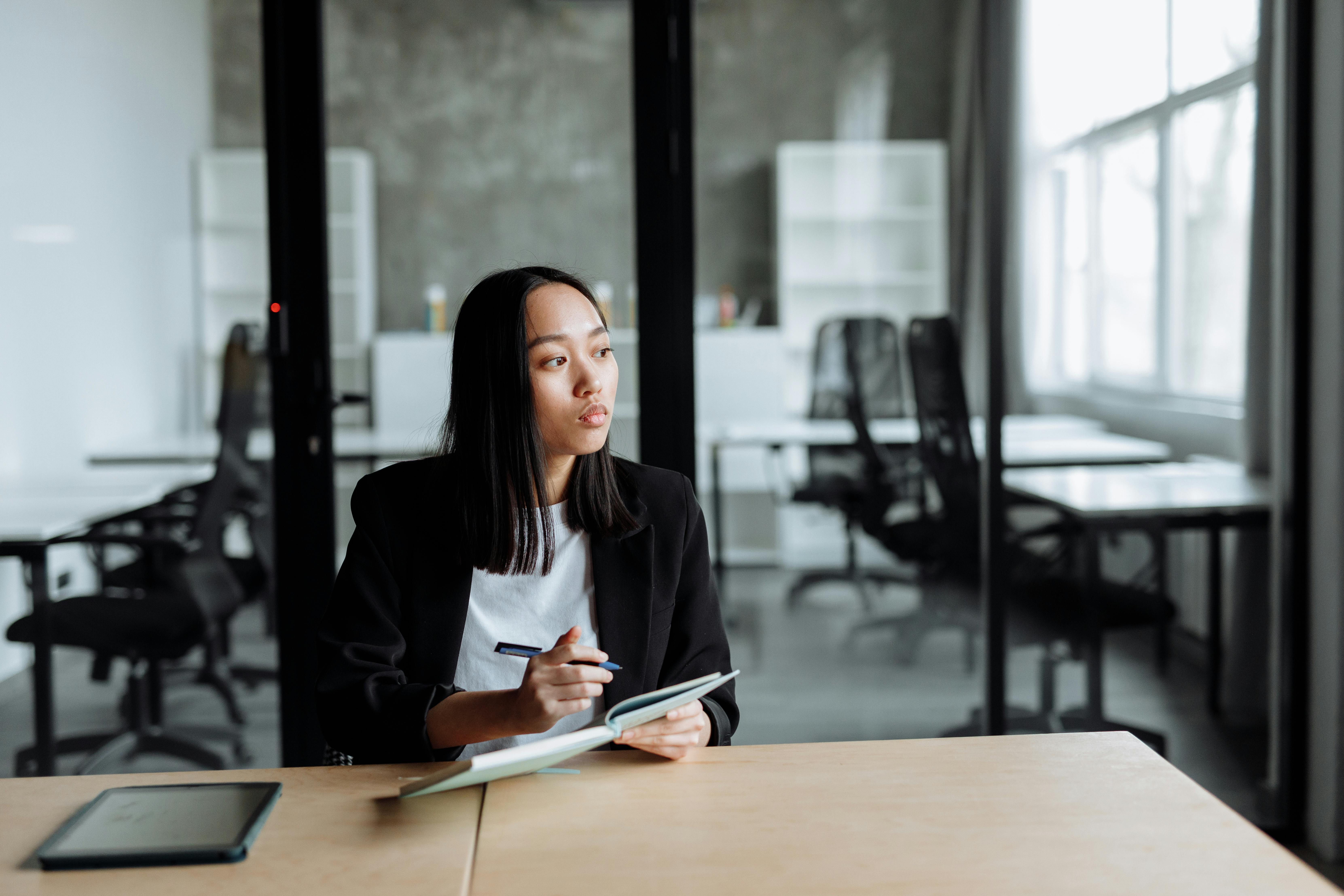 A young woman in a black blazer sitting at a desk, writing in a notebook in a modern office setting.