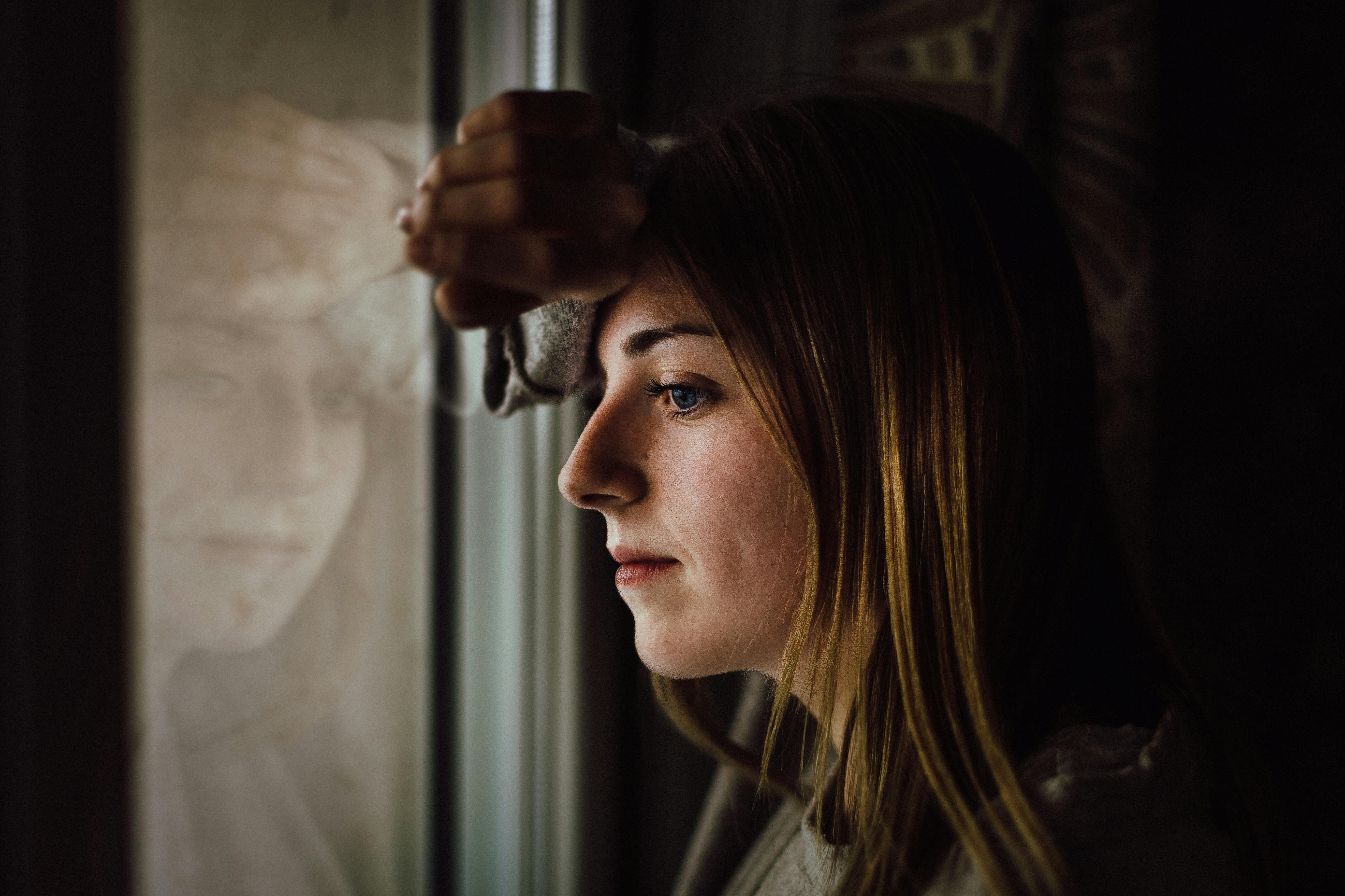 A woman with long hair resting her forearm on a window, gazing thoughtfully outside.