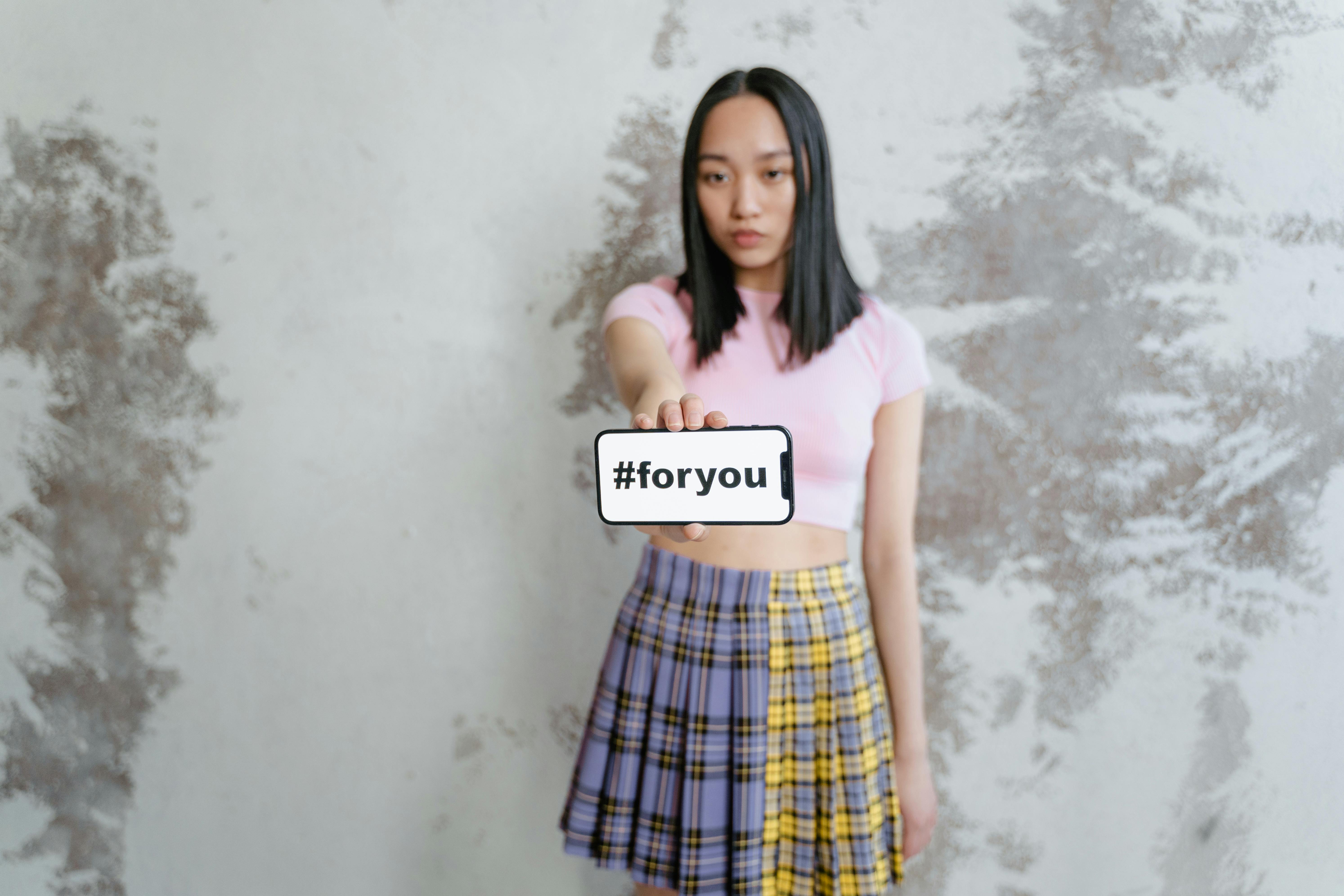 A young woman holding a phone displaying the hashtag #foryou, wearing a pink top and a plaid skirt.