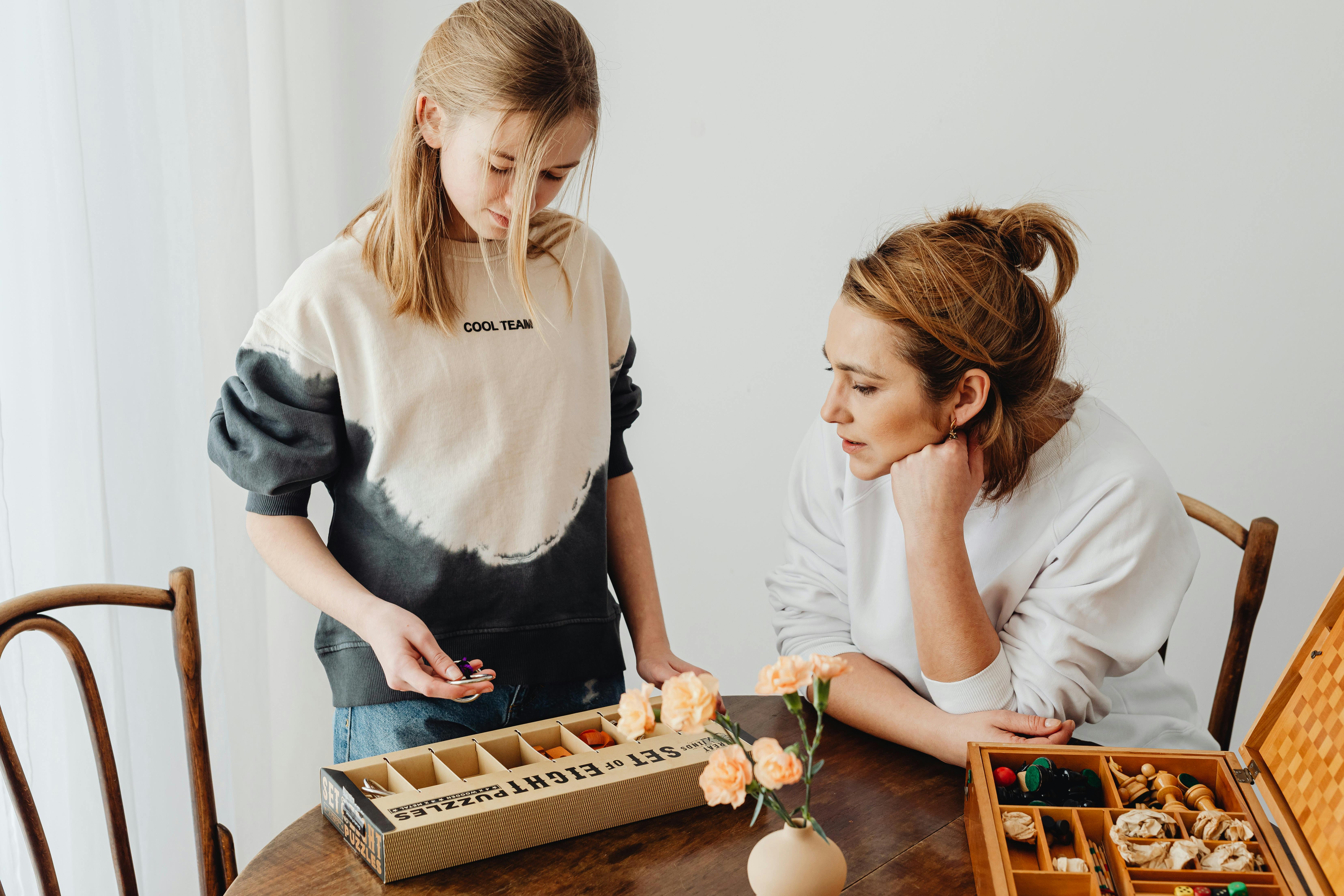 A girl and a woman engaged in a puzzle activity at a table with flowers in a vase.