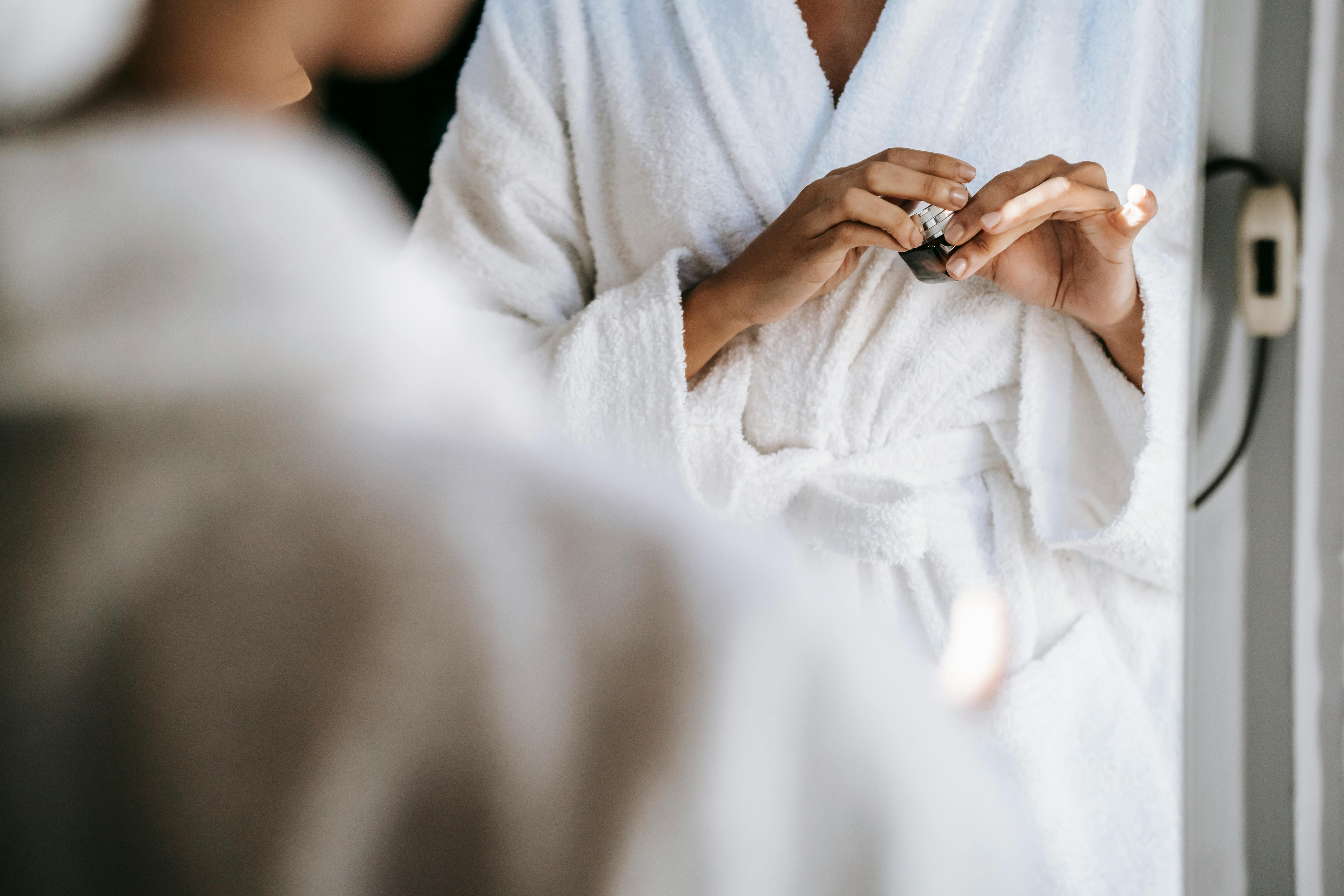 Person in a white bathrobe applying skincare product in front of a mirror