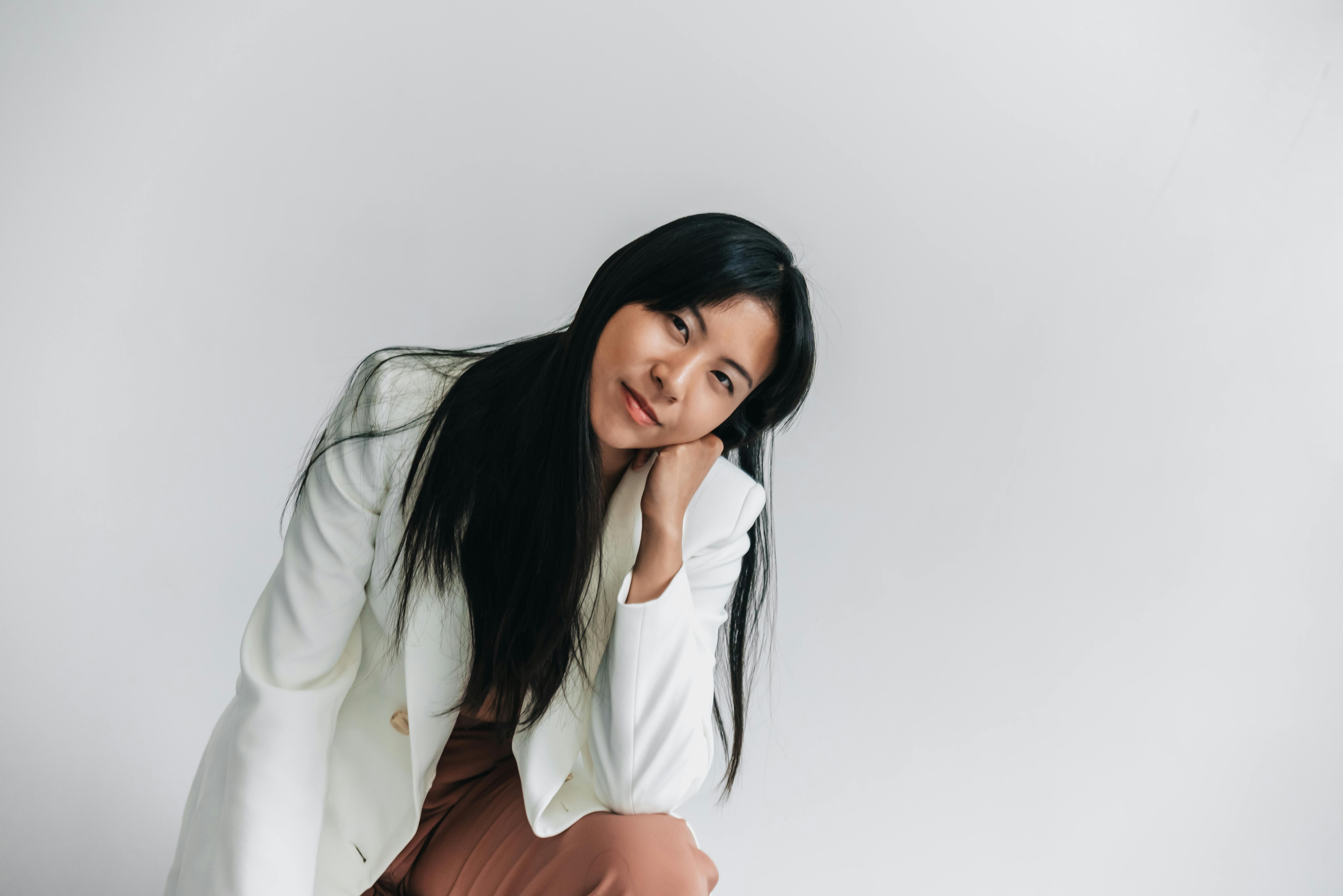 A woman in a white blazer, sitting and resting her chin on her hand with a thoughtful expression.