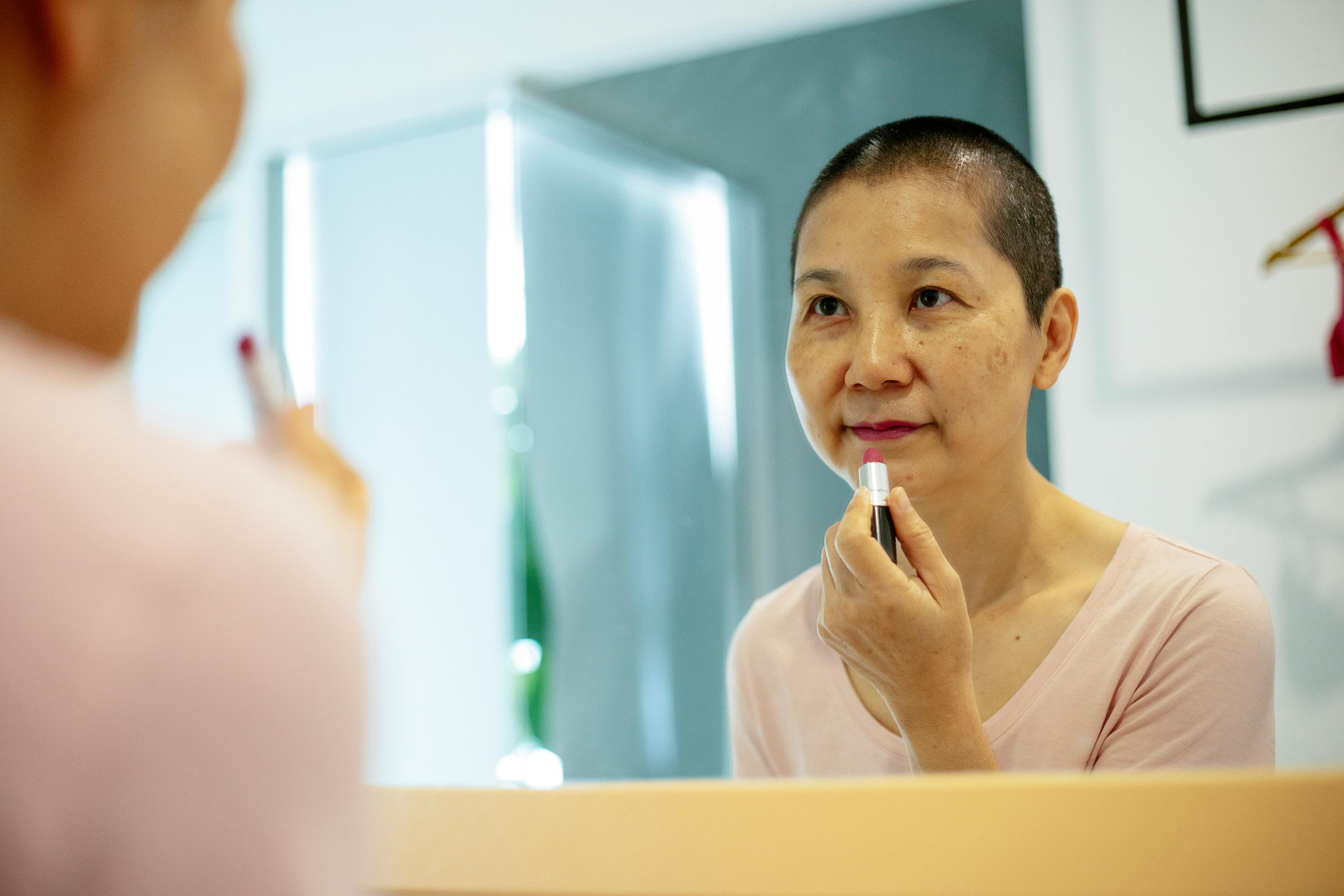 A woman with a shaved head applying lipstick in front of a mirror.