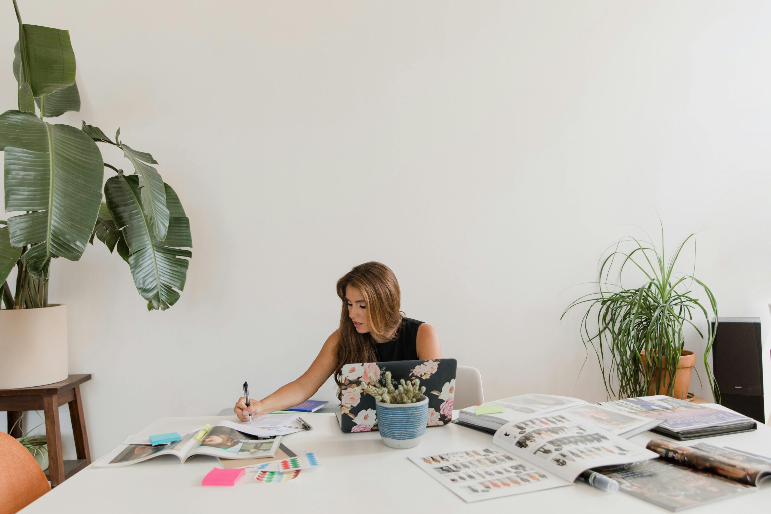 A woman sitting at a desk with a laptop, writing notes, surrounded by magazines and a potted plant.