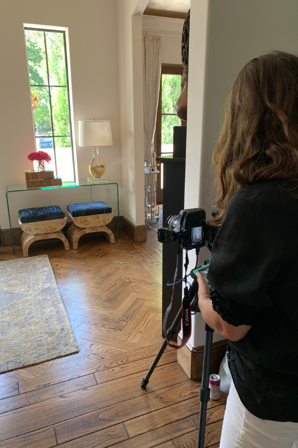 a woman photographing an interior vignette with two stools, flowers, and a vase with a gold base in front of a long window