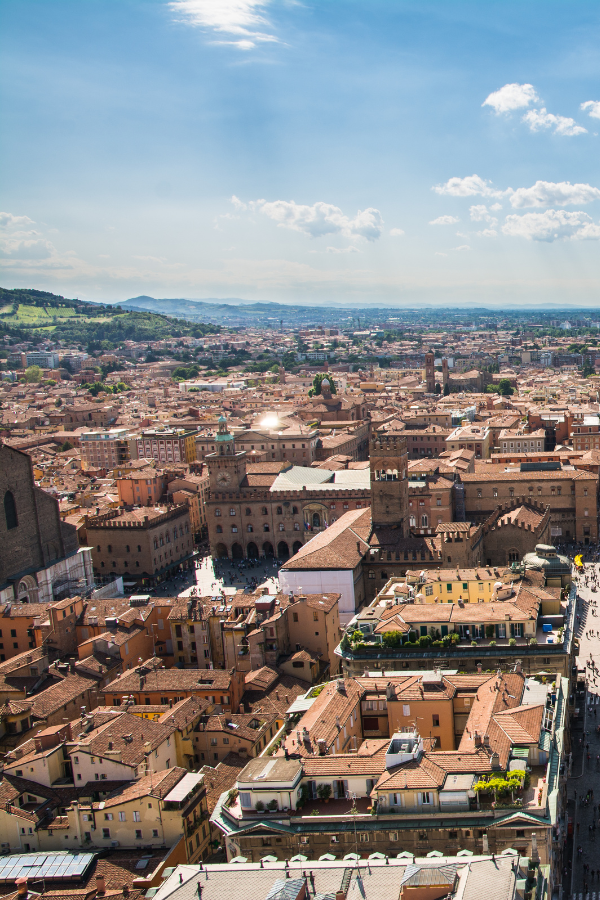Elevated view over Bologna’s terracotta rooftops, with a large historic brick basilica and a green-domed church in the foreground.