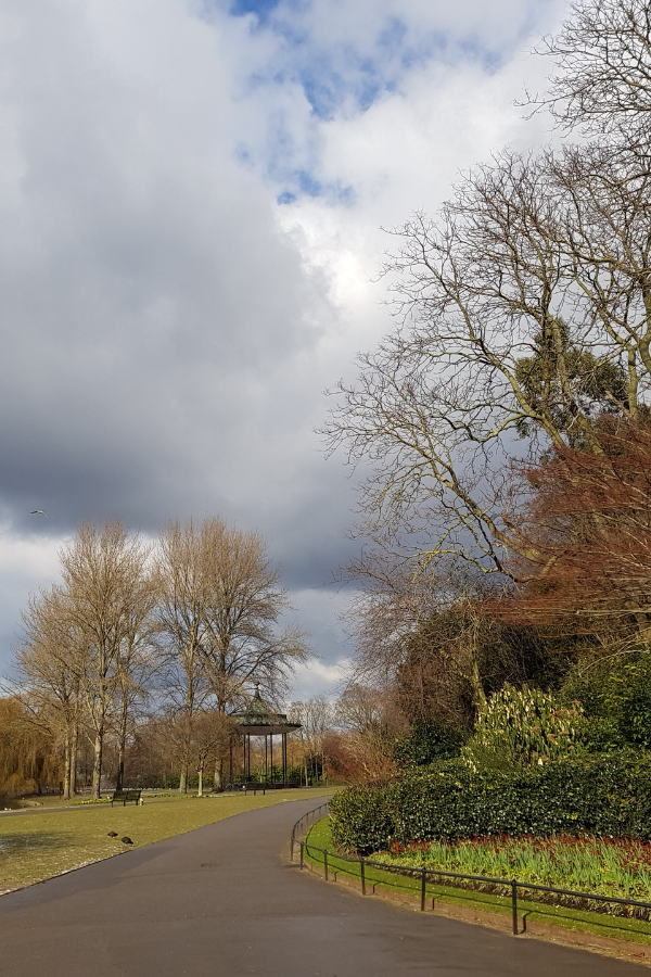 A curving path through Regent’s Park beside landscaped flower beds and leafless trees, beneath dramatic grey-and-white clouds.