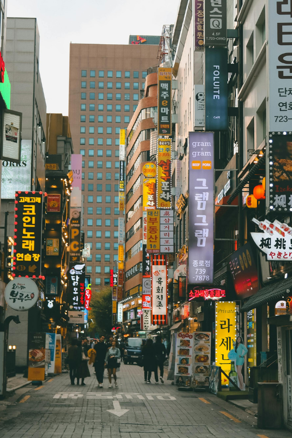 A busy pedestrian street in Seoul lined with restaurants, shops, and colorful Korean signs rising between tall buildings.