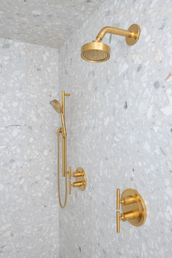Brass rainfall shower and handheld shower fixtures mounted on pale terrazzo-style stone tiles.